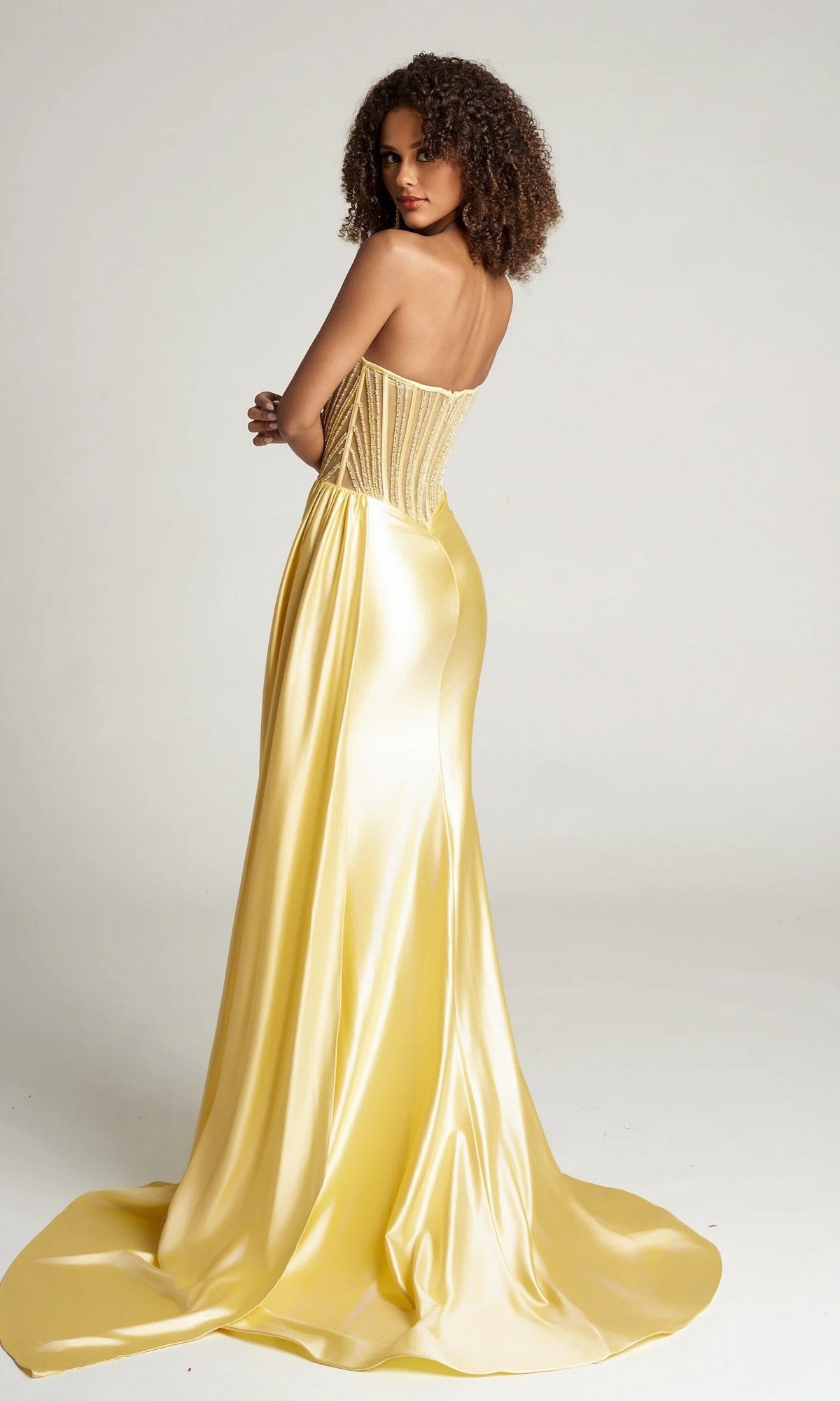A woman in the Portia and Scarlett PS26056 long formal dress with a strapless fitted bodice and flowing skirt stands facing away, glancing over her shoulder against a plain background.